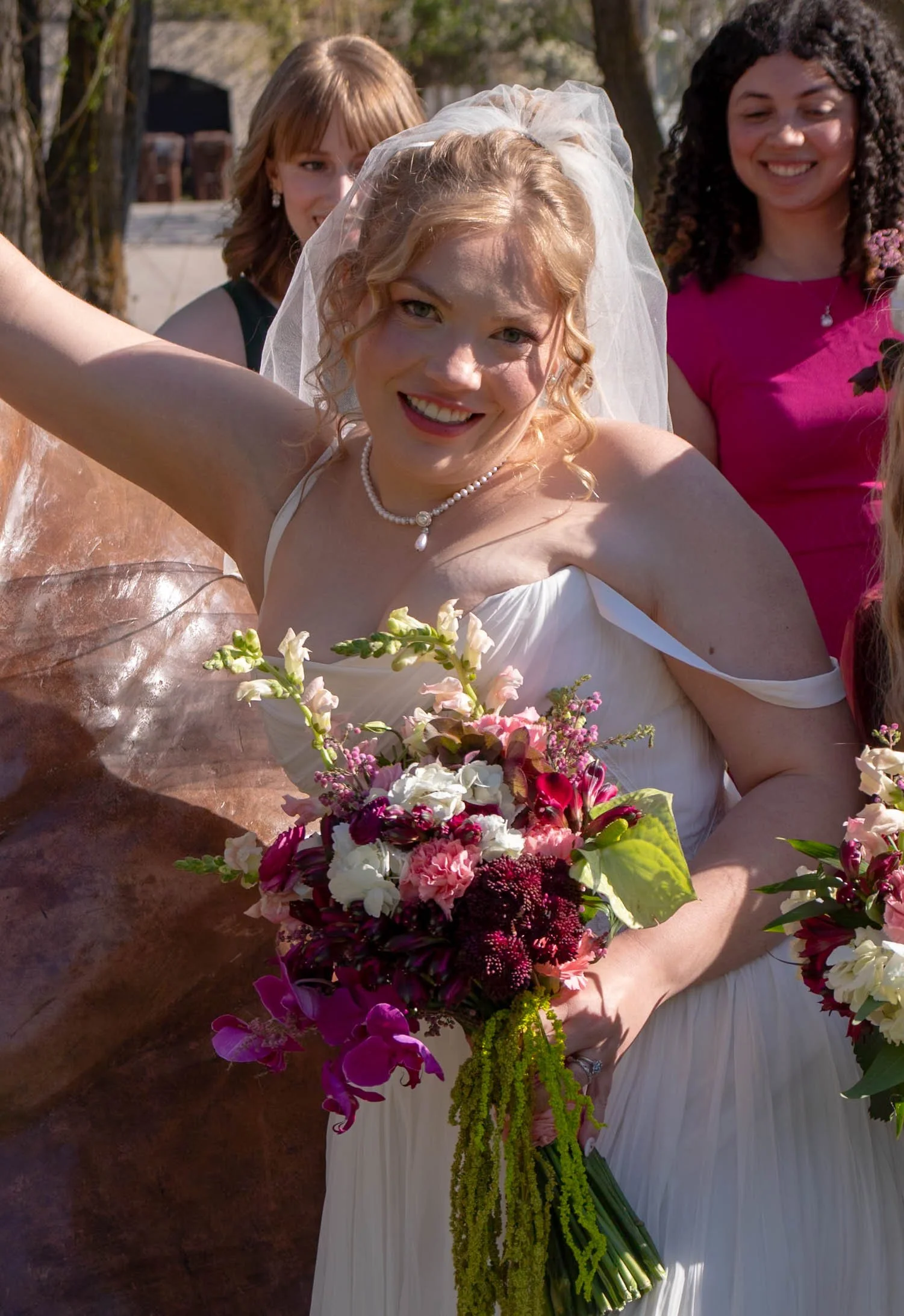 A bride in a white dress wearing a veil and pearl necklace holding a large bouquet of pink, white, and purple flowers, smiling outdoors with three women in the background.