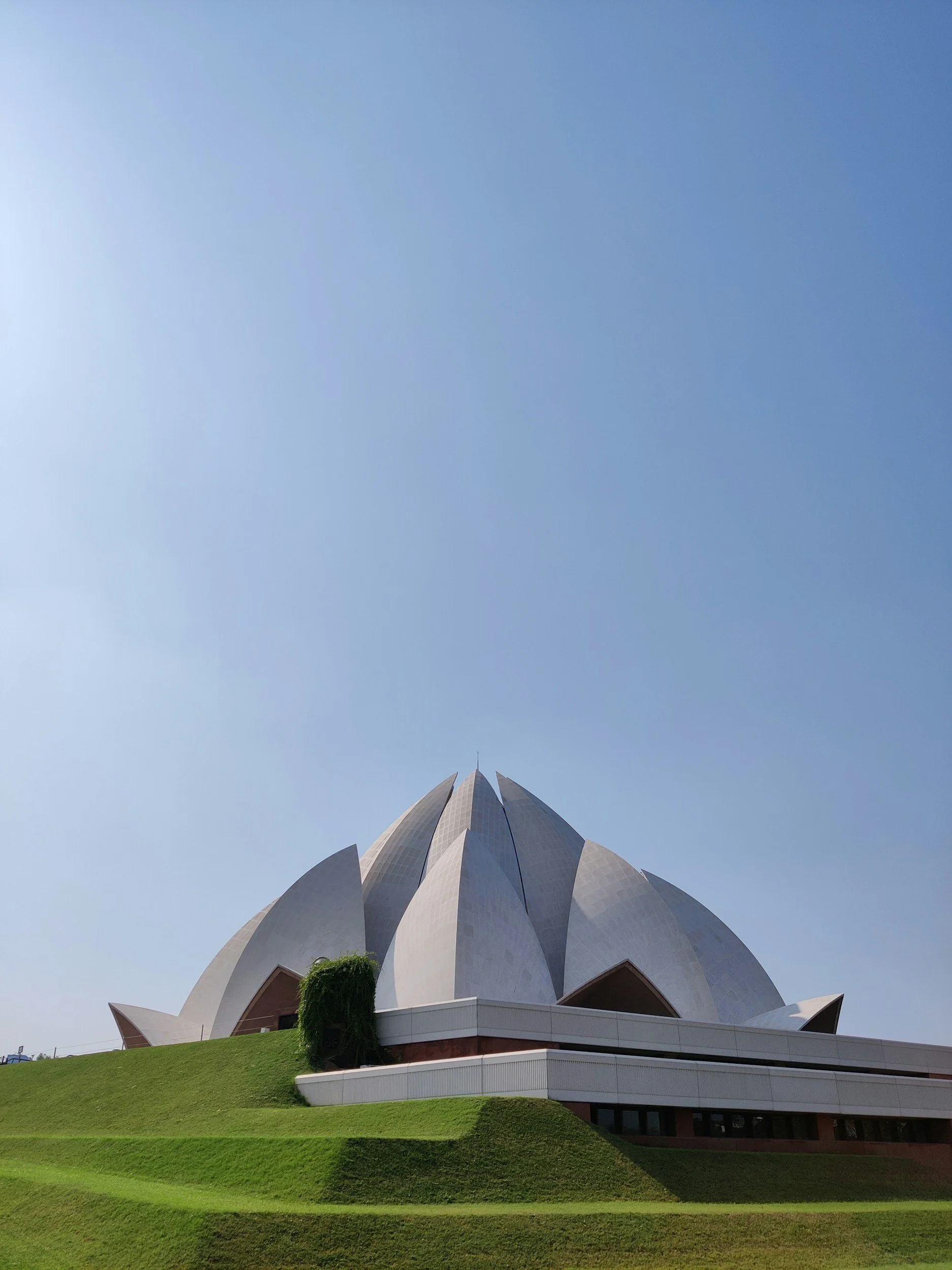 View of the Lotus Temple in Delhi, India, with white petal-like structures rising from a lush green lawn against a clear blue sky.