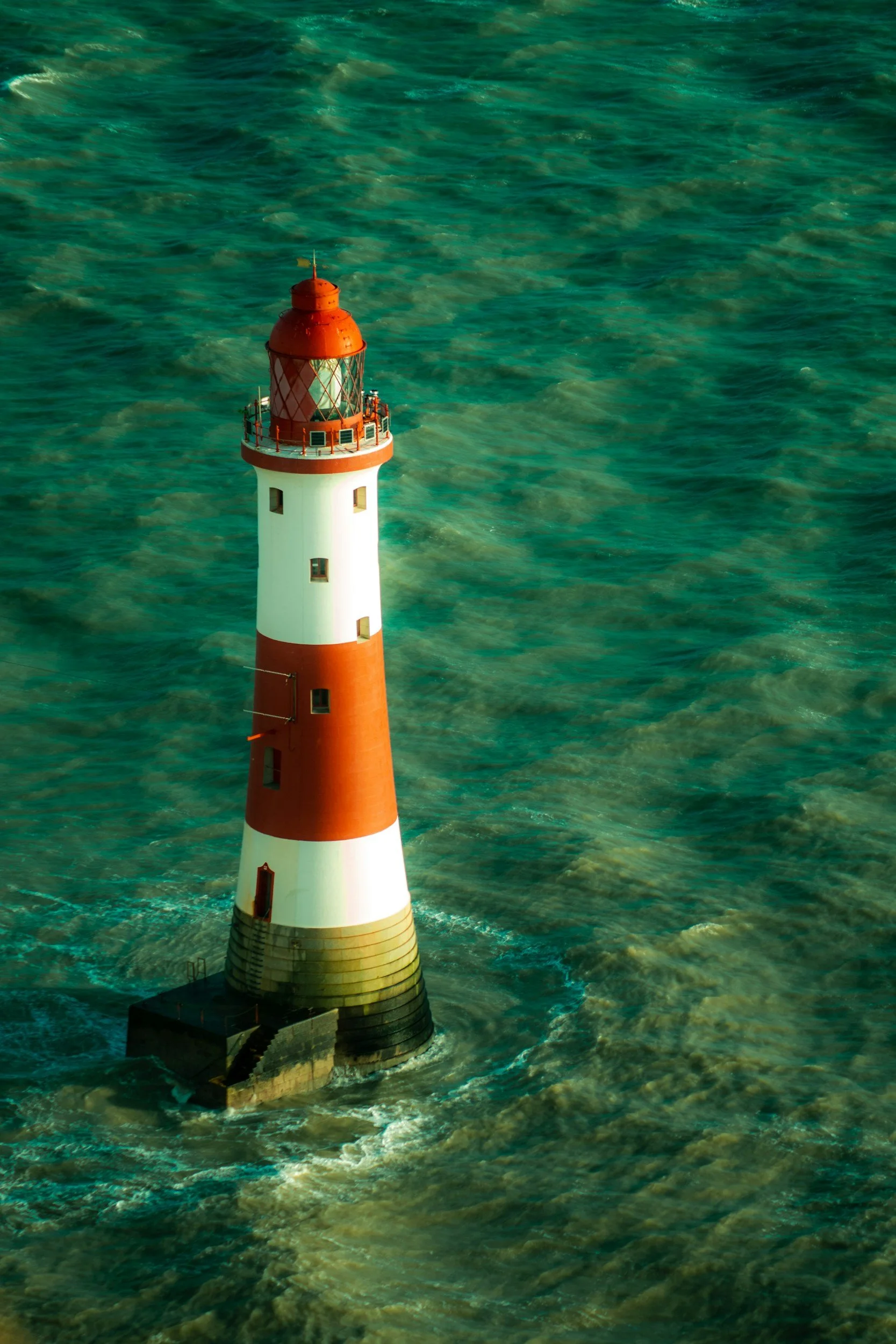 A tall white lighthouse with red stripes and a red top, surrounded by greenish water with waves.