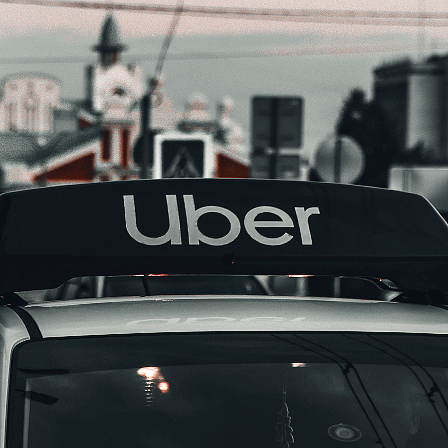 Uber vehicle with a sign on the roof in an urban area, background includes buildings and utility wires.