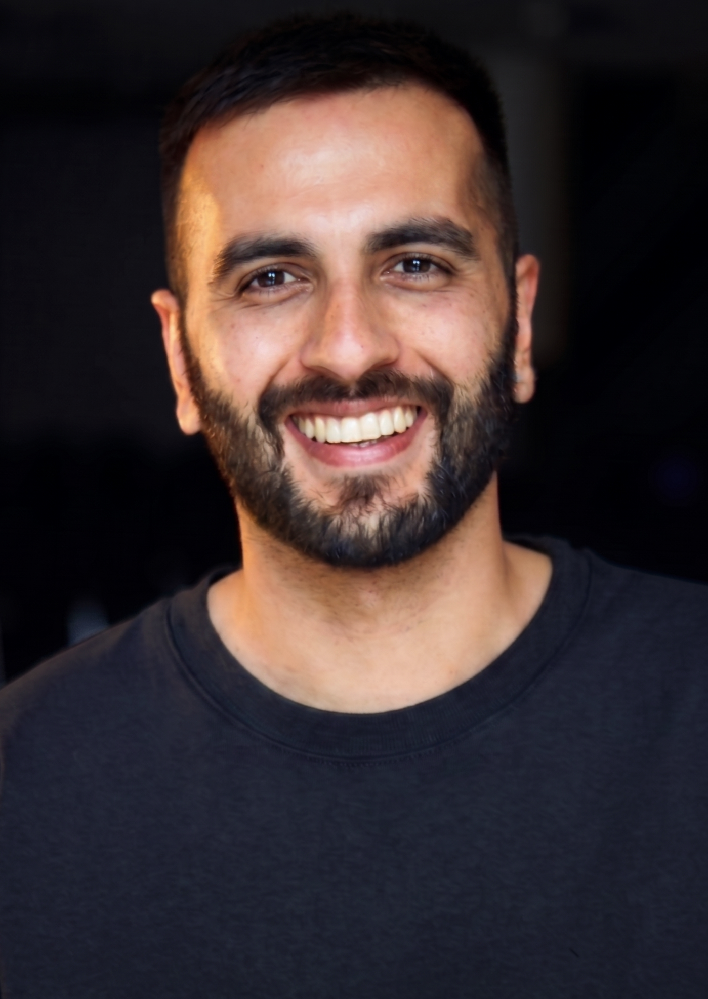 Close-up of a smiling man with dark hair and a beard, wearing a black T-shirt, against a dark background.