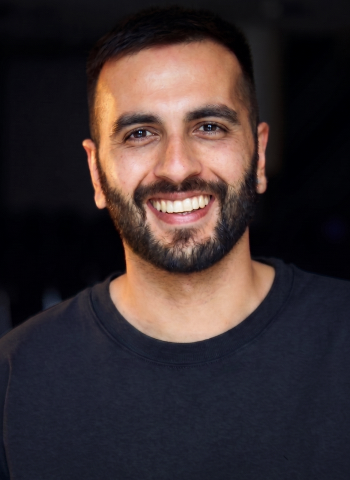 Close-up of a smiling man with dark hair and a beard, wearing a black t-shirt, against a dark background.