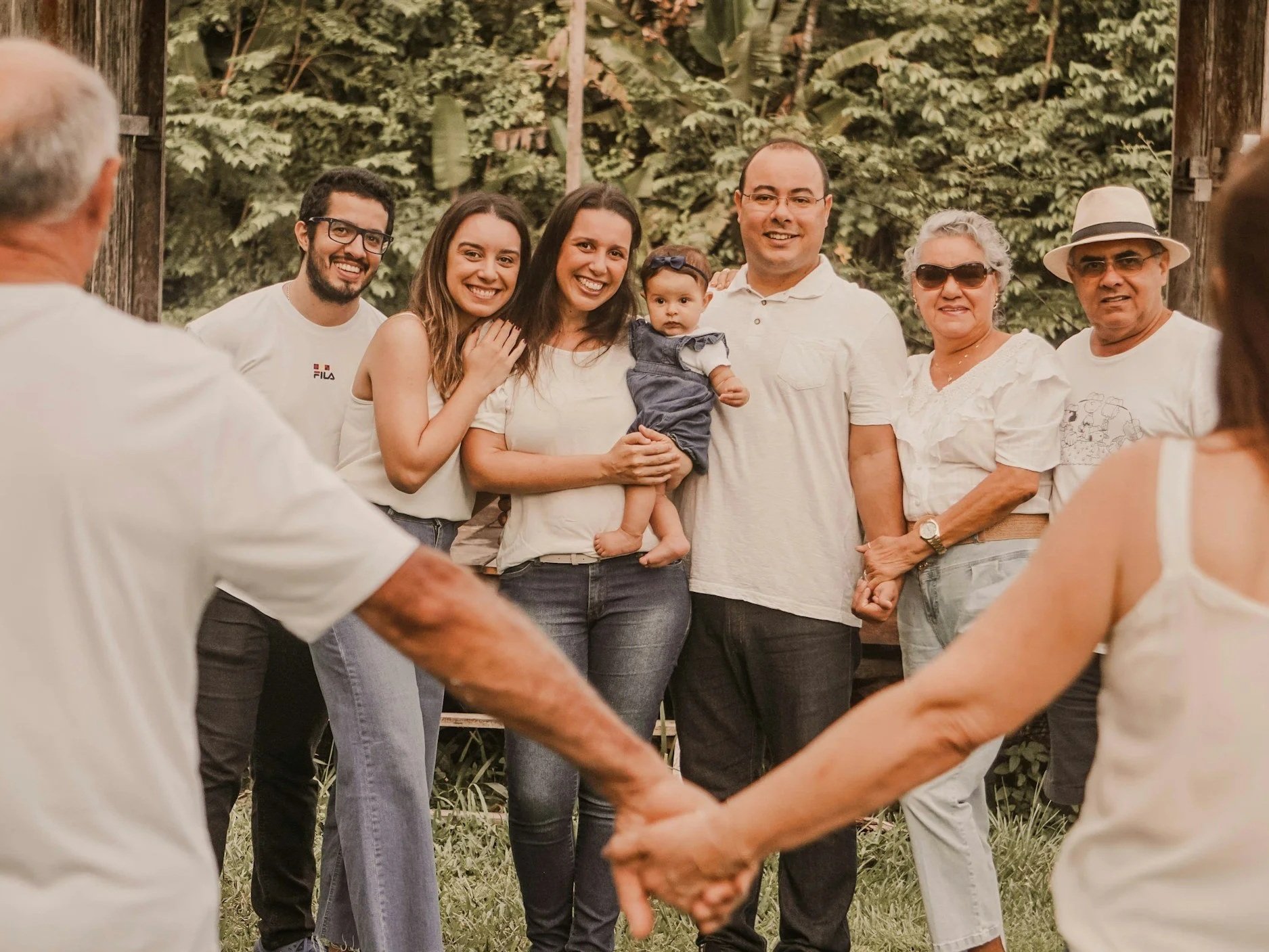 A group of people holding hands in a circle outdoors, with some also embracing in the center, smiling and enjoying a gathering in a natural setting with trees and greenery.
