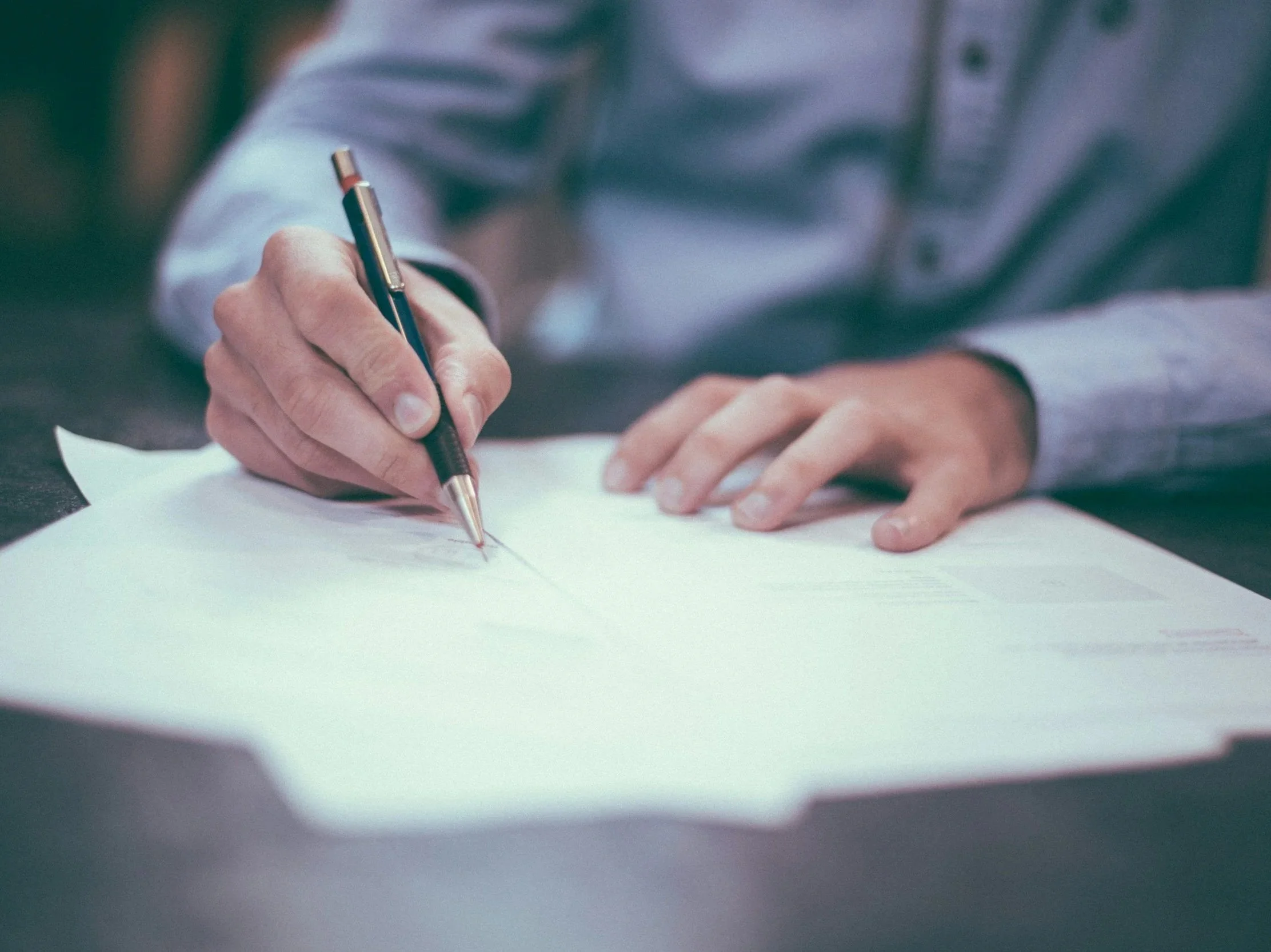 Close-up of a person's hand holding a pen and writing on a sheet of paper on a dark surface.