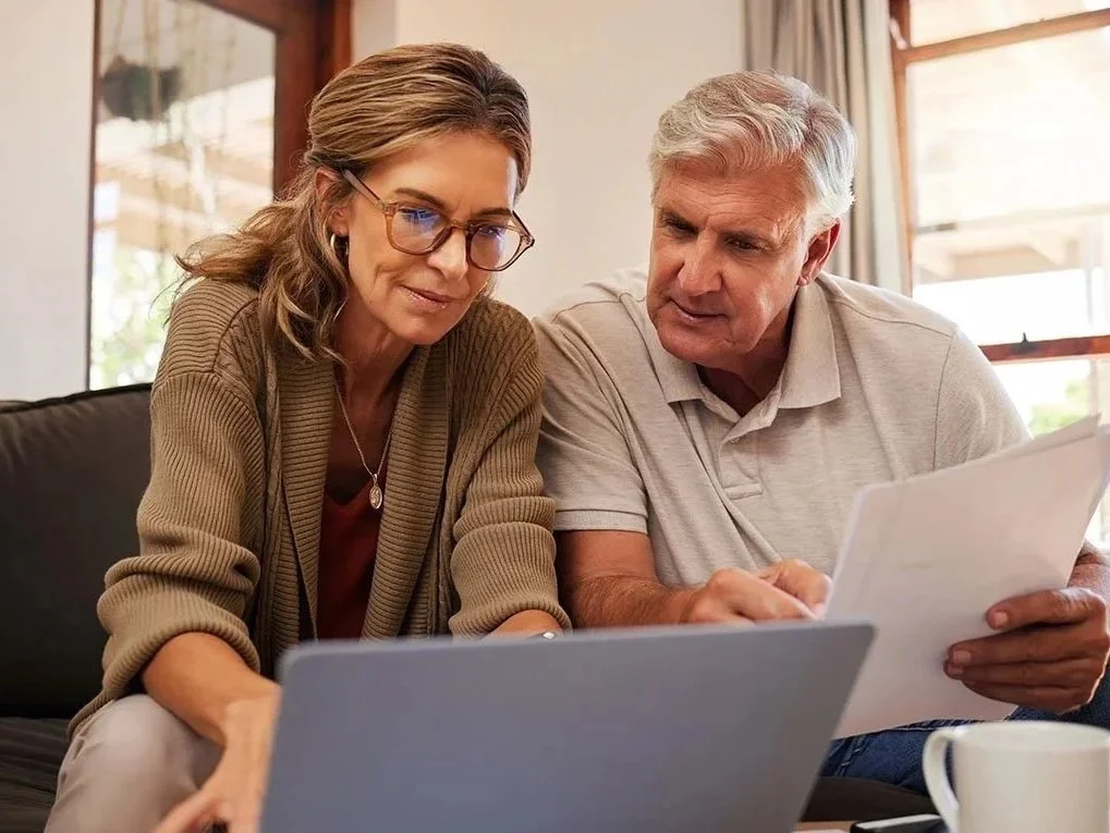 A senior man and woman sitting on a couch looking at documents and a laptop, with a coffee mug nearby in a bright room with large windows.