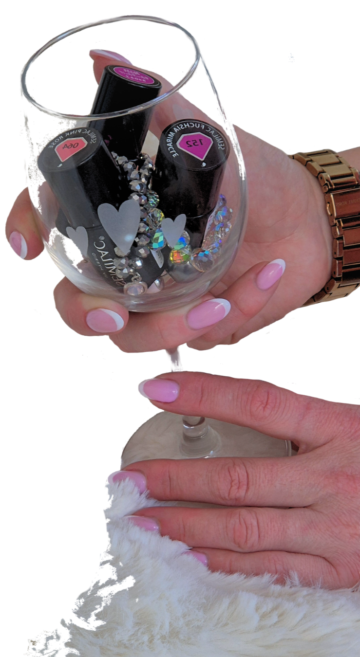 A feminine hand holding a wineglass containing two gel nail polishes. The hand has classic, soft pink and white, French tip nails and is wearing a gold watch. The other hand is resting on the bottom of the glass, on a furry white blanket.