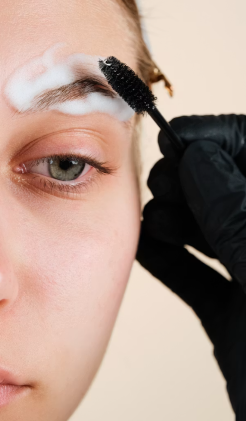 A person applying white cream or makeup to their eyebrow with a black spoolie brush, held by a gloved hand.