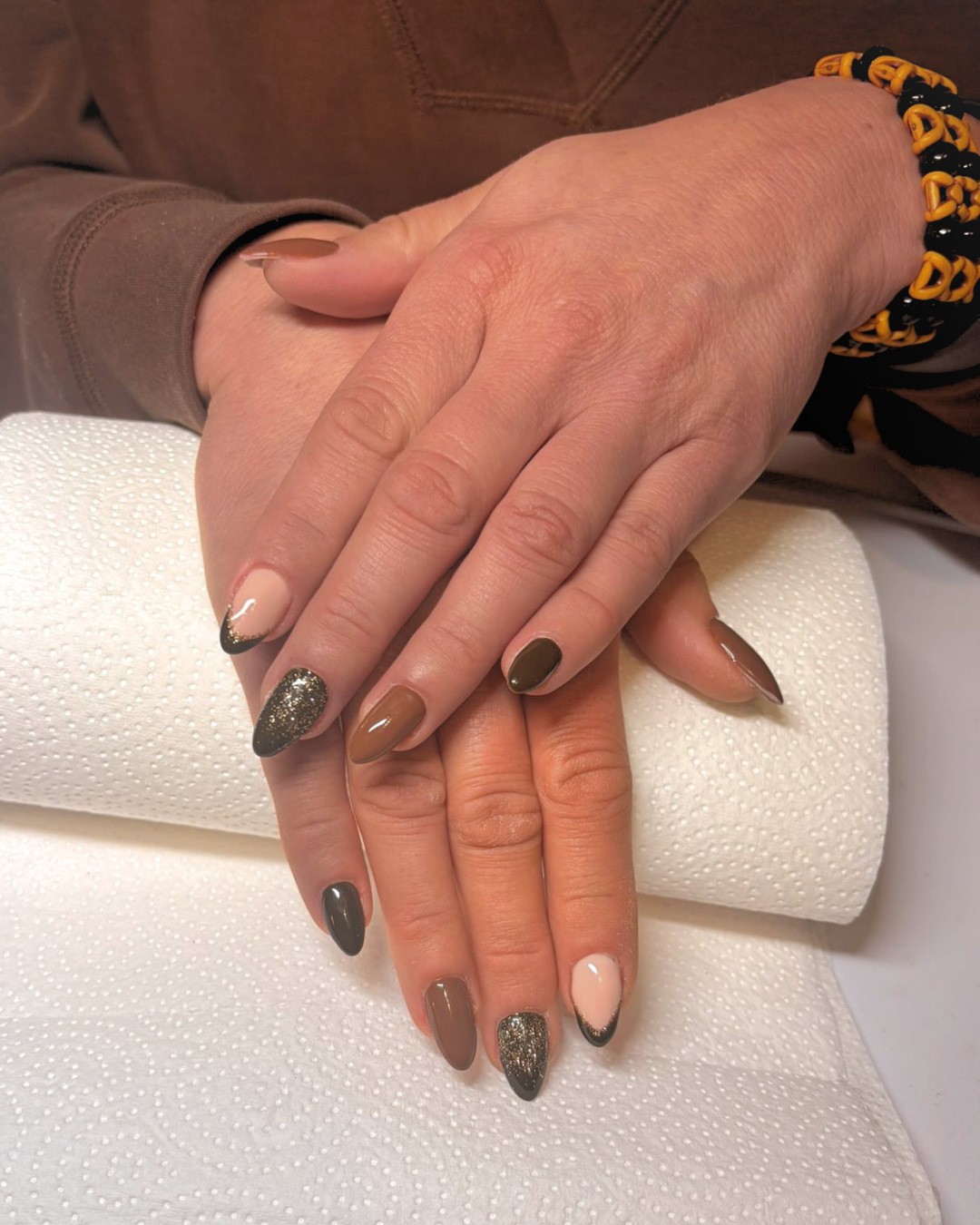 Close-up of hands with long almond-shaped gel nail extensions, featuring brown, beige and glittery black gel polish, resting on a paper towel roll. The person is wearing a brown long-sleeve shirt and a yellow and black beaded bracelet.