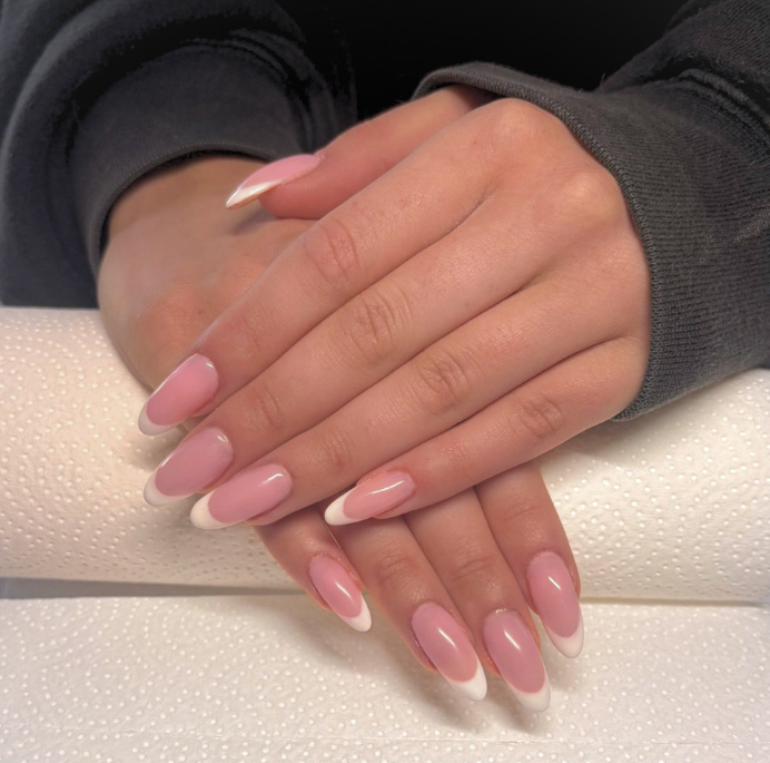 Close-up of hands with gel nail extensions featuring a natural pink base and white french tips, resting on a textured white surface.