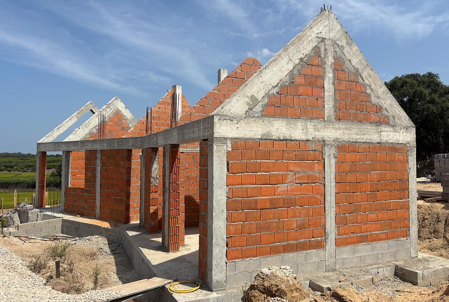 Under construction brick and concrete building with a triangular roof shape, set against a blue sky with some clouds, surrounded by open land.