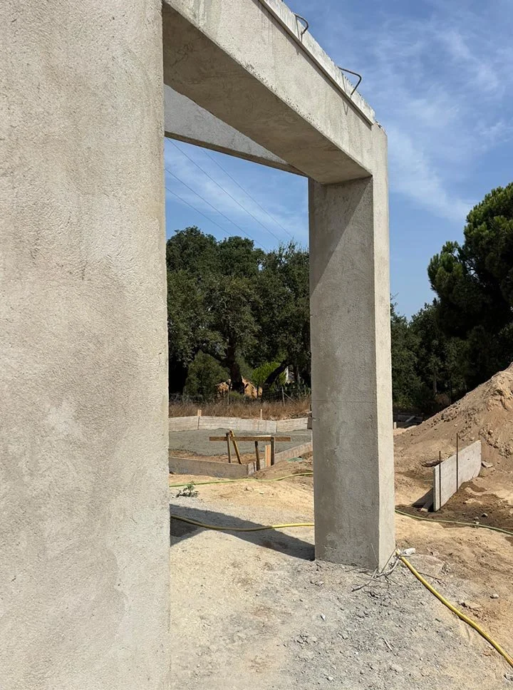 Part of a building under construction with a concrete support column and a partial concrete wall, with construction tools and dirt surrounding the area.