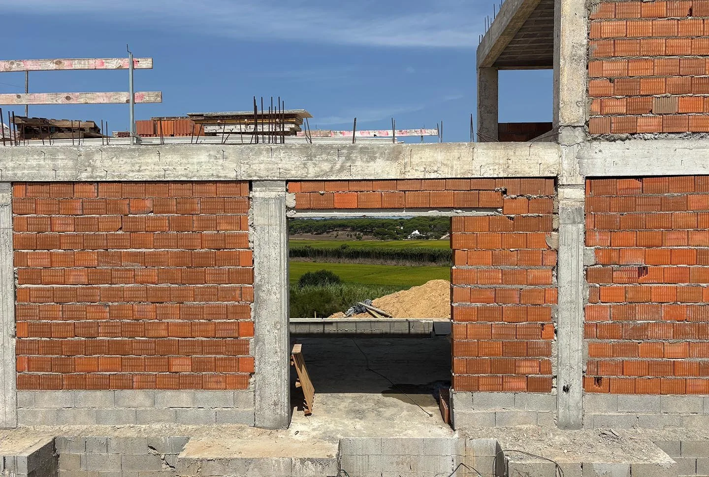 Partially constructed brick building with open window, overlooking green fields and distant trees under blue sky.