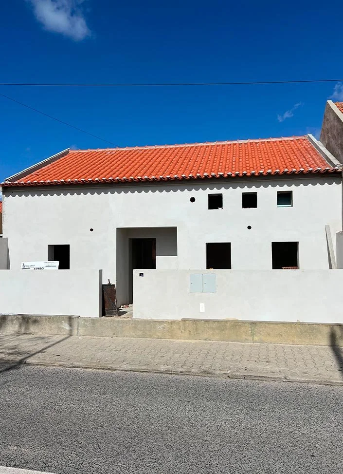 A white house with a red tiled roof, multiple small window openings, and a front gate, under a blue sky.