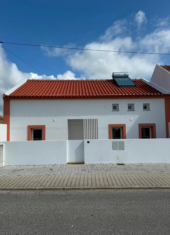 A modern house with a red-tile roof, white walls, and small windows, situated behind a white fence with a paved sidewalk in front.