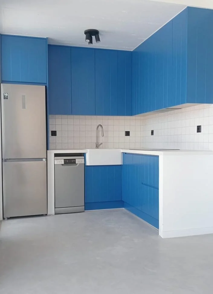 A modern kitchen with blue cabinets, a white farmhouse sink, a dishwasher, a stainless steel refrigerator, and a white tiled backsplash.