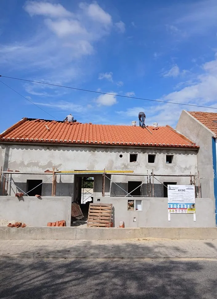 Under construction house with a red tile roof, scaffolding, and workers on the roof during daytime.