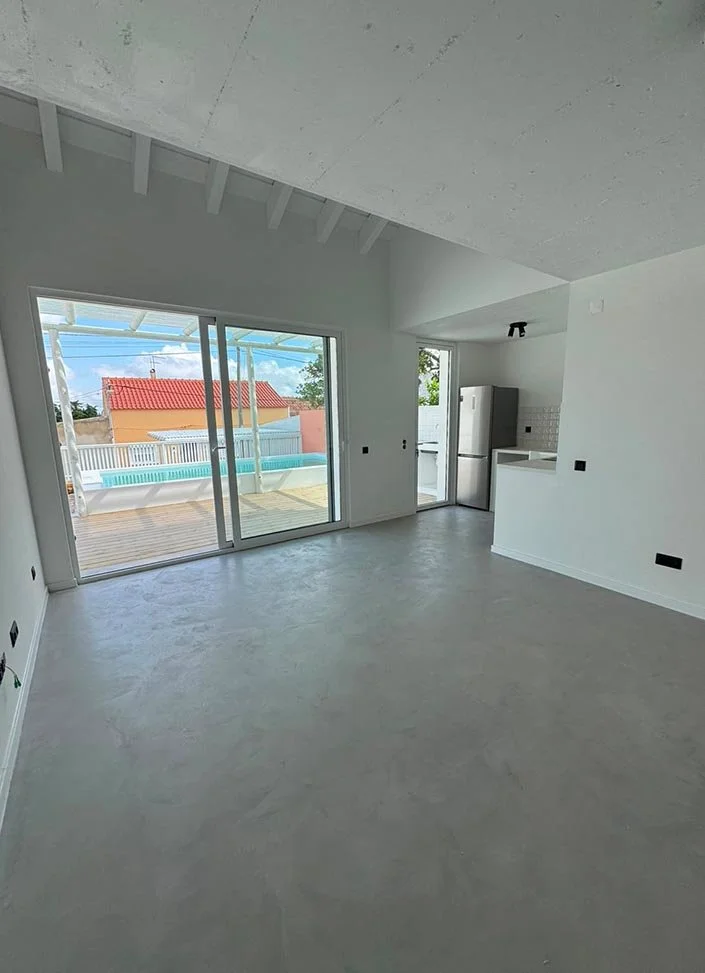 Empty modern living room with large glass sliding doors leading to an outdoor patio, and a small kitchen area with a stainless steel refrigerator.