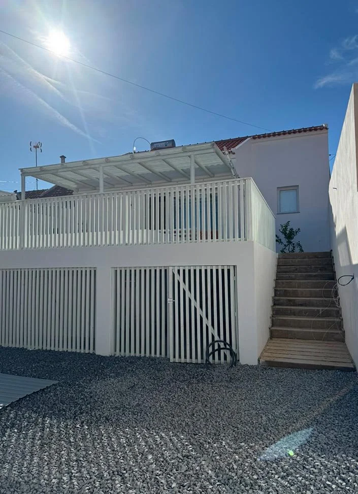 White residential building with a wooden staircase, balcony with metal railing, clear blue sky, and the sun shining brightly.