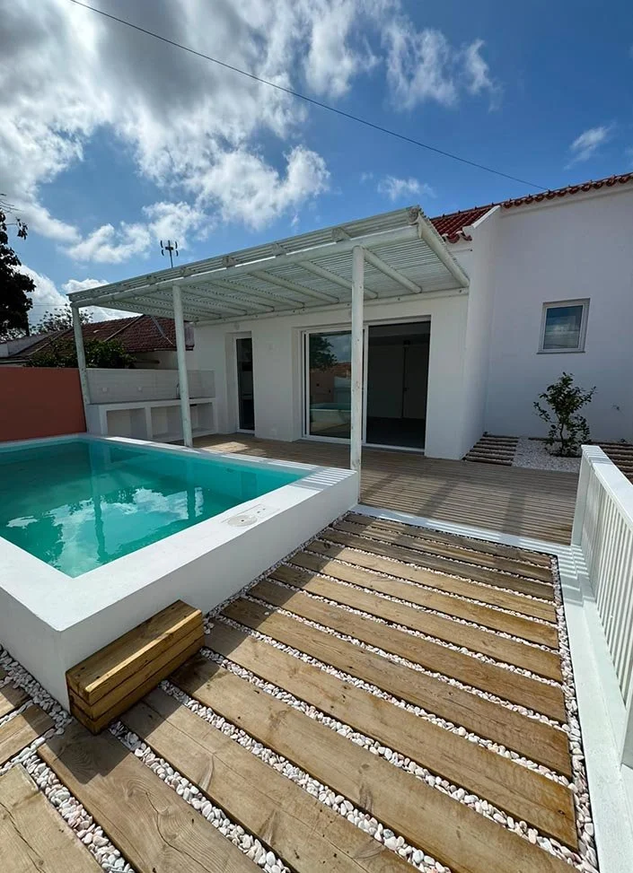Modern backyard with a small rectangular pool, wooden deck, and white patio rails, attached to a white house under a partly cloudy sky.
