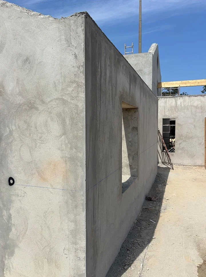 A concrete wall with a square window opening under construction, with scaffolding and construction materials around, and a clear blue sky above.