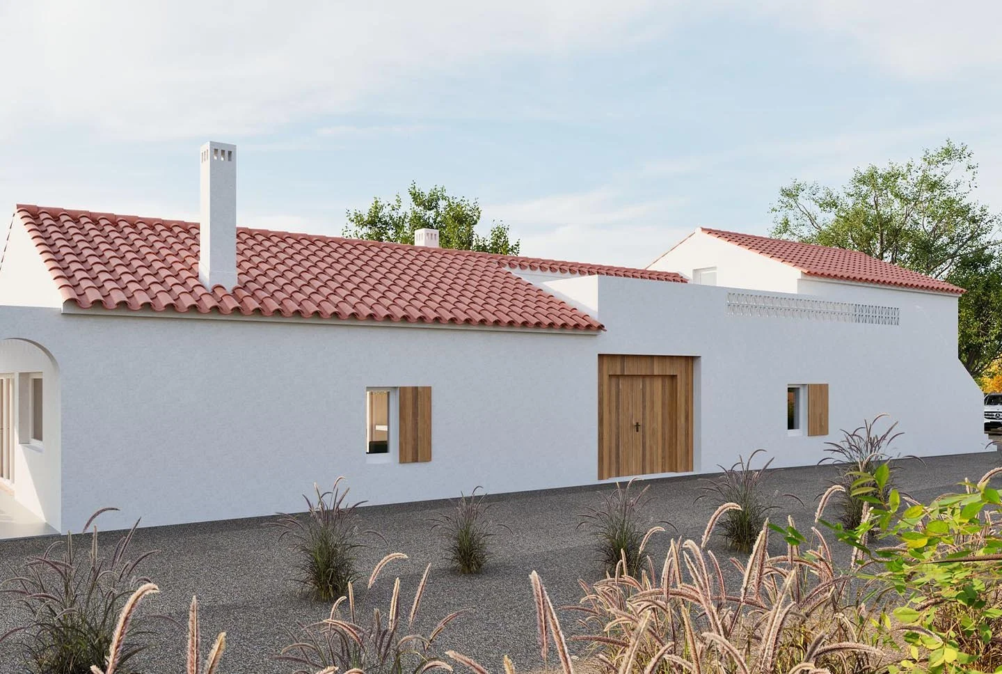 Modern white house with red tile roof, wooden doors and window shutters, and a gravel yard with ornamental grasses, set against a partly cloudy sky and trees in the background.
