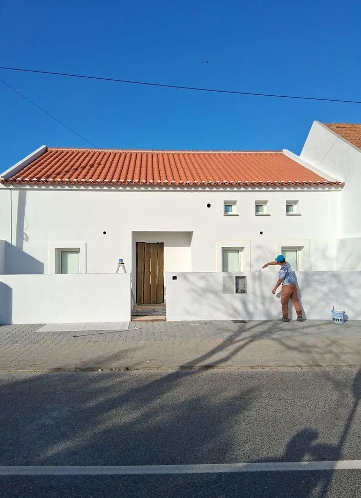 A person standing outside a white house with a red-tiled roof, painting the wall. There is a bucket of paint on the ground, and the house has small rectangular windows. The sky is clear and blue.