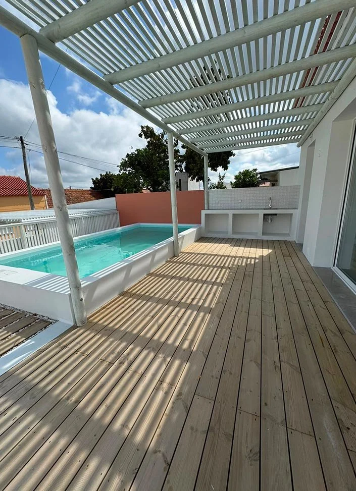 Backyard patio with a small in-ground pool, wooden deck, and a white pergola. The scene includes a partial view of a sink area with an outdoor faucet, a brick wall, trees, and neighboring houses in the background.