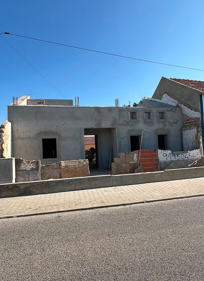 A house under construction with unfinished walls and no windows, located along a paved street with a curb and a sidewalk, under a clear blue sky.