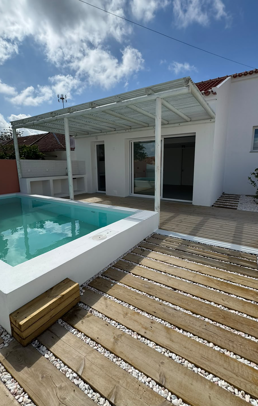 A modern backyard patio with a swimming pool, wooden decking, and a white house with sliding glass doors. There is a small shaded area with a slatted roof and a privacy wall. The sky is partly cloudy.