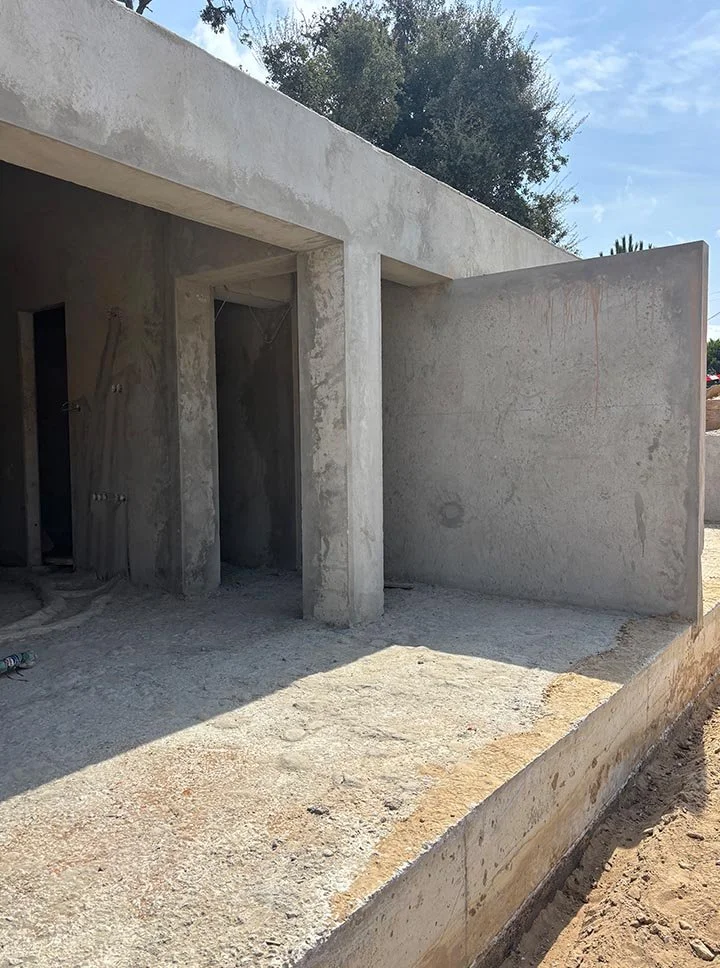 An unfinished concrete building under construction, with an open doorway and a side wall, surrounded by dirt and construction materials.