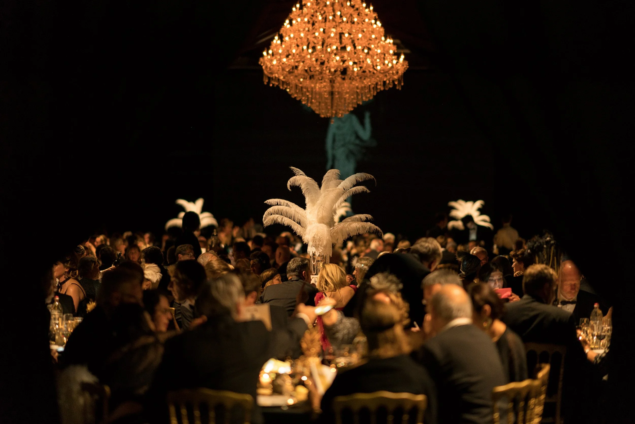 Une grande salle avec une multitude de personnes assises à des tables, décorée avec un grand chandelier en cristal suspendu au plafond, au centre d'une décoration avec des plumes blanches.