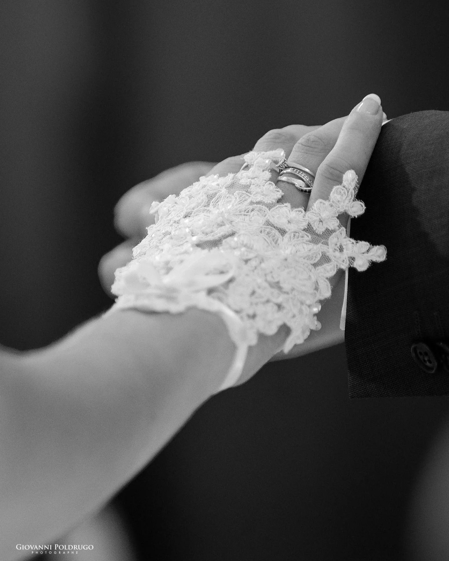 Une main d'une femme portant une robe en dentelle qui touche la main d'un homme en costume noir, dans un contexte de mariage ou de cérémonie élégante.