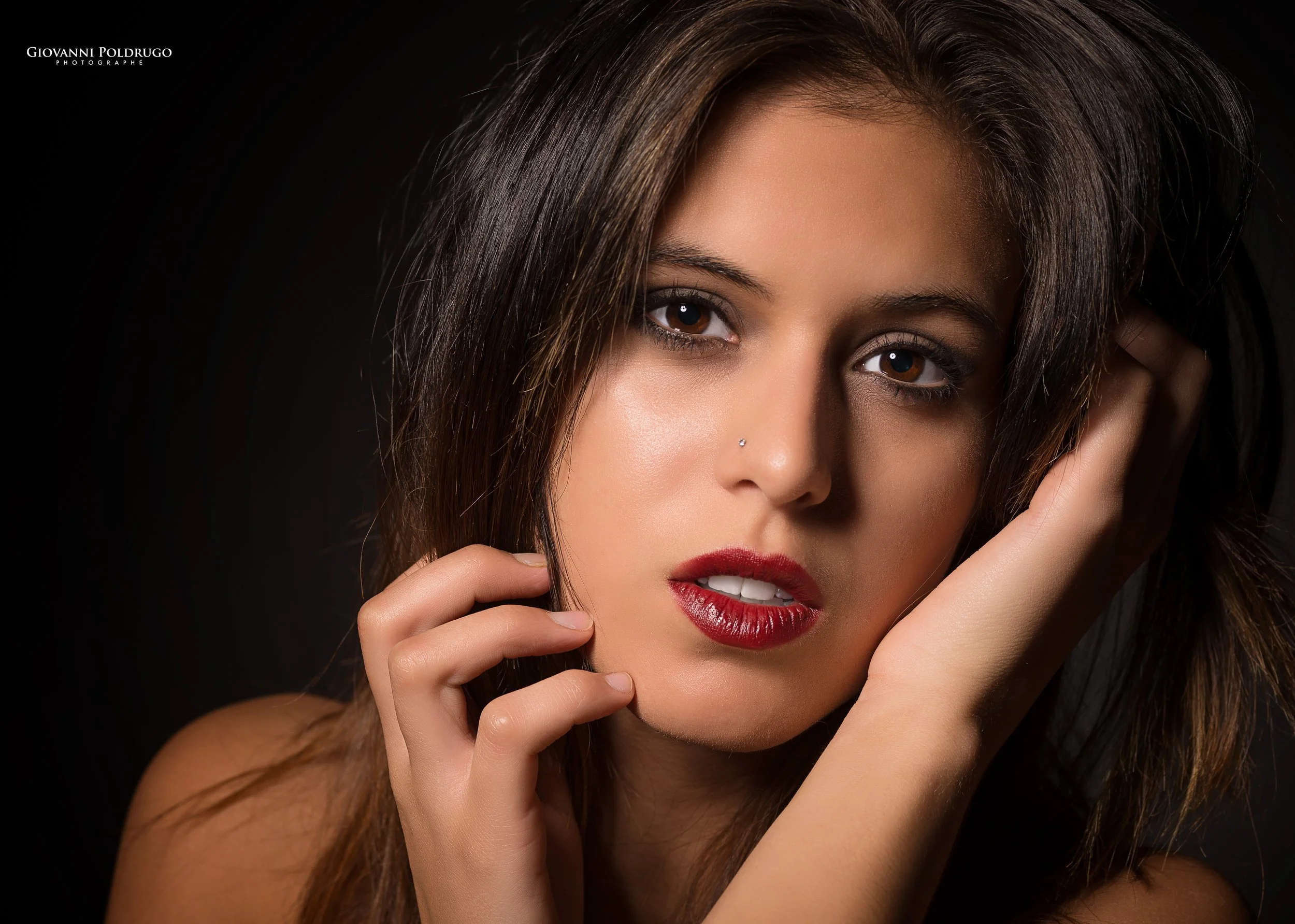 Portrait d'une femme avec des cheveux bruns, maquillage soigné, portant du rouge à lèvres rouge, touchant son visage avec ses doigts, fond noir.