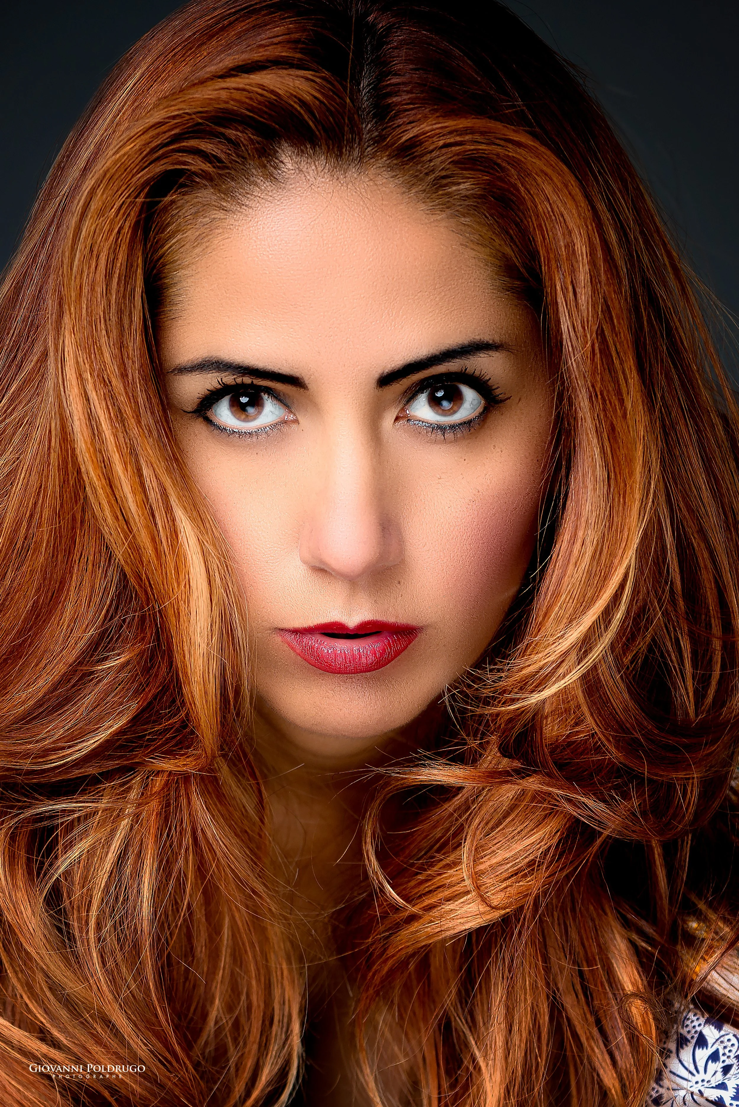 Un gros plan du visage d'une femme aux cheveux roux bouclés, avec un maquillage sophistiqué mettant en valeur ses yeux et ses lèvres. Fonds sombre.