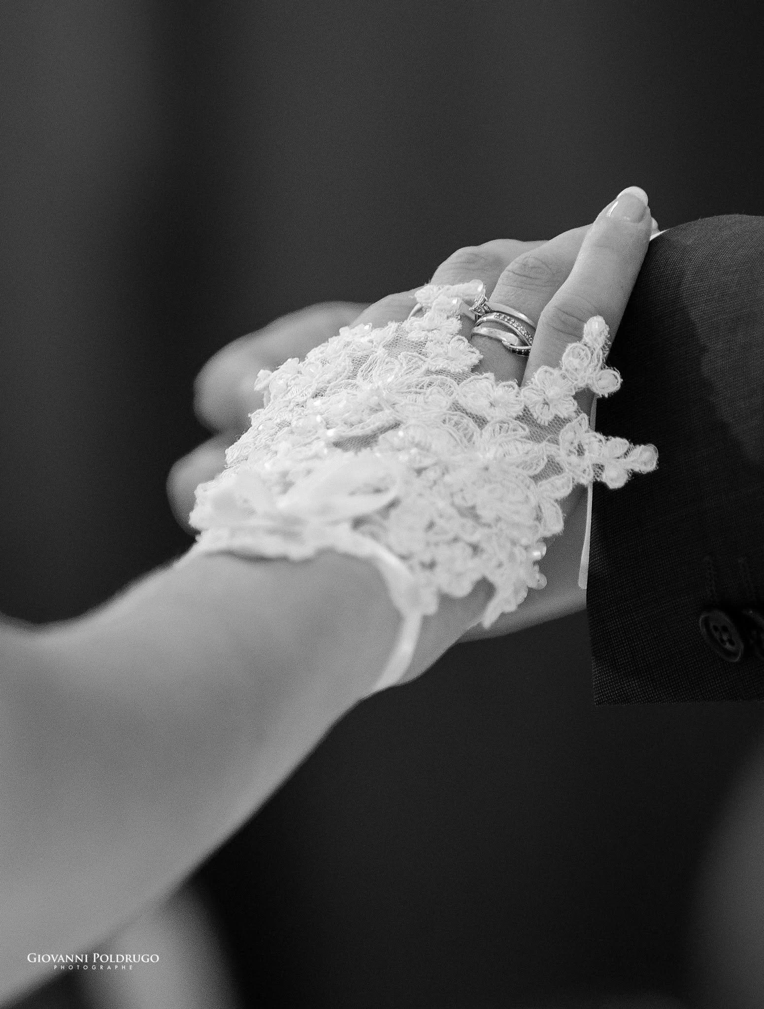 Une main d'une femme portant une bague et une manche en dentelle, tenant la main d'une personne vêtue d'un costume noir. Photo en noir et blanc.