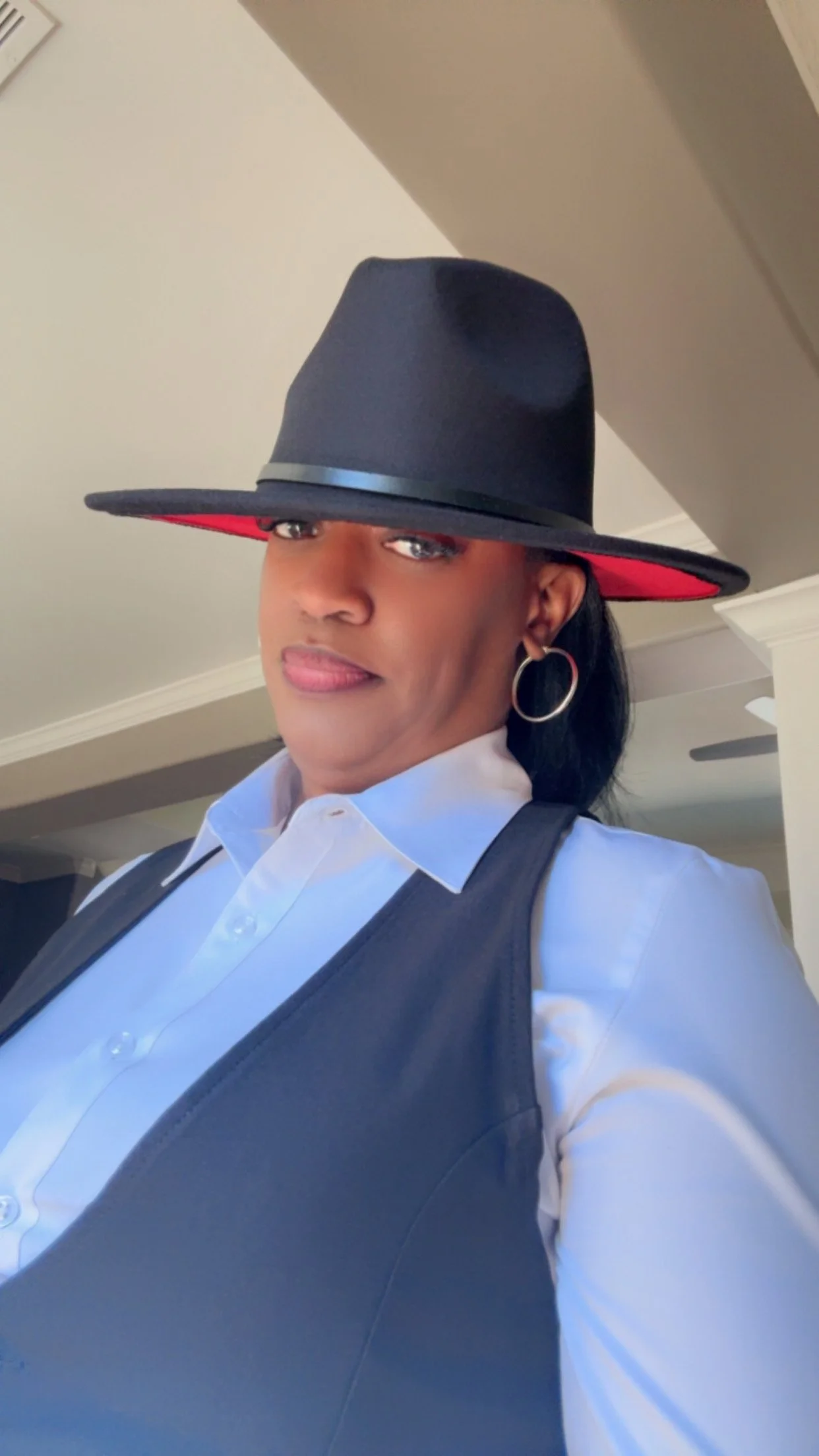 A woman wearing a white dress shirt, a black vest, and a black hat with a red underside, standing indoors.