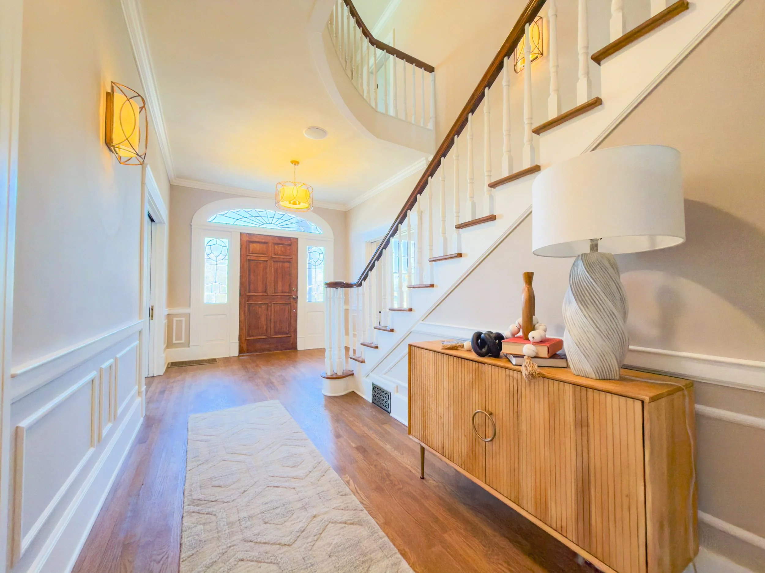 Interior view of a spacious entryway with wooden flooring, a front door with decorative glass, a staircase with white spindles and wooden handrails, and a wooden sideboard with a lamp and decorative items in Pulaski, TN.
