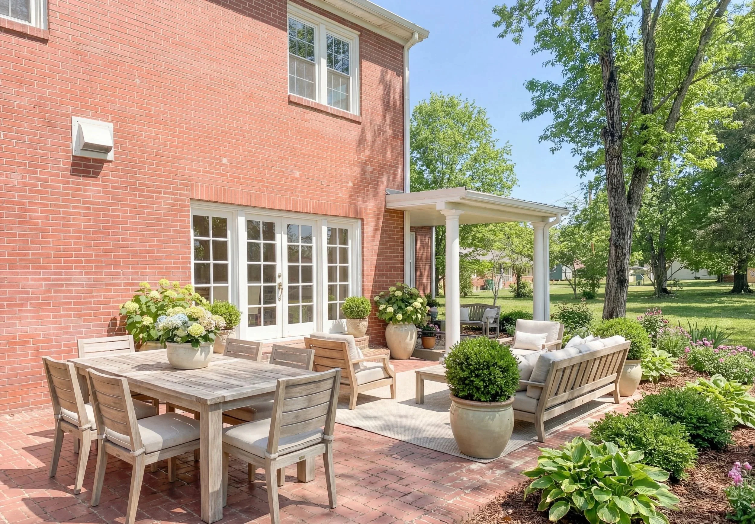 An outdoor patio area with a brick house in the background, featuring a wooden dining table with six chairs, potted plants with flowers, and a seating area with cushioned furniture under a small porch with white columns. The patio is surrounded by lush green trees and garden beds with plants and flowers.