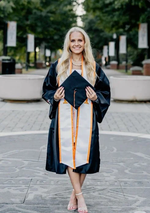A young woman in a graduation gown holding a cap at the University of Tennessee Southern, standing outdoors on a paved area with trees and signs in the background.