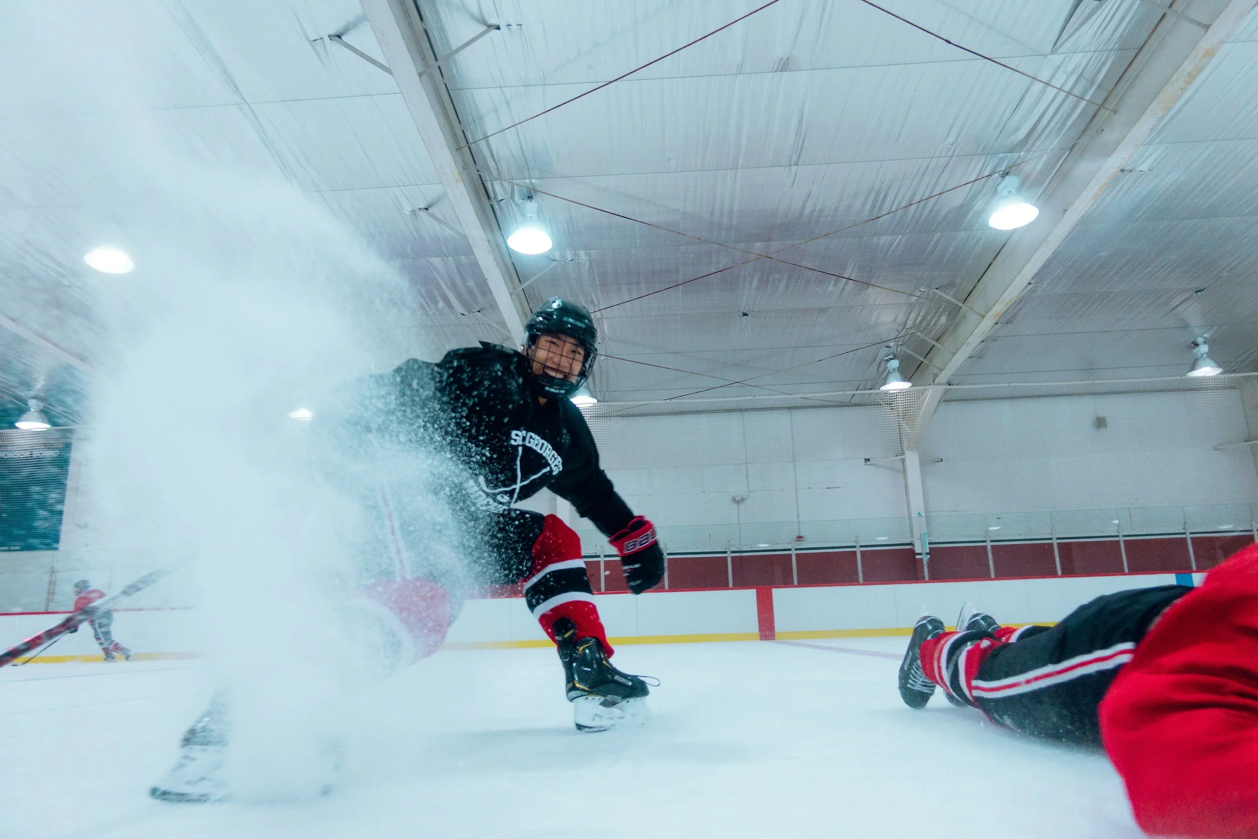 An ice hockey game with players in action, one player in black and red gear falling on the ice and another in a black jersey skating nearby, inside an indoor rink with bright overhead lighting.
