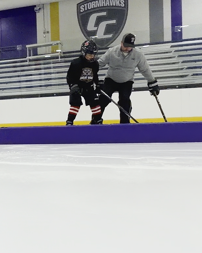 An adult hockey coach and a youth hockey player on an ice rink, practicing hockey. The adult is helping the child with their footwork in a skating drill. In the background, there is a sign that says "Stormhawks" with a logo.