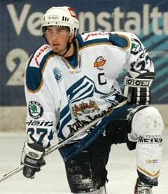 Ice hockey player in a white uniform with blue and gold accents, wearing a helmet and holding a hockey stick on the ice.