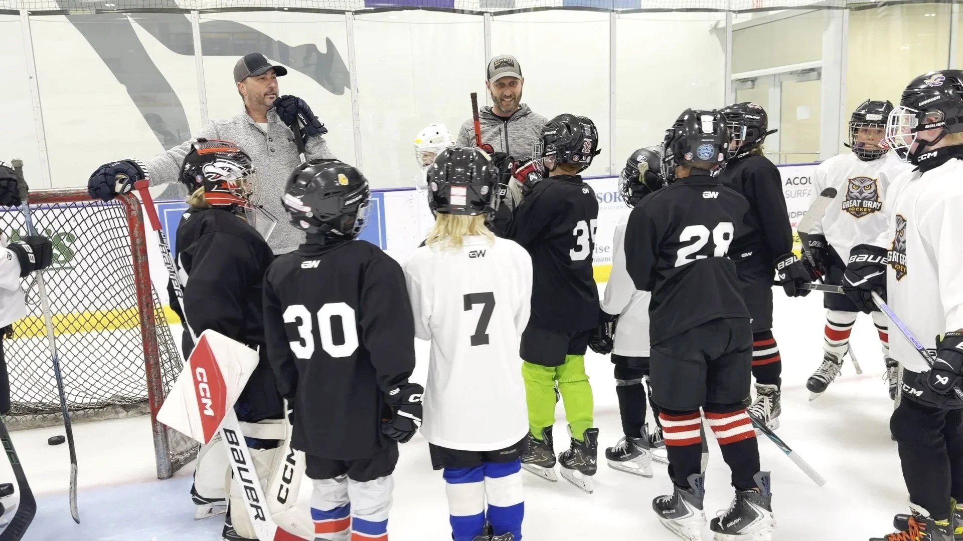 Hockey coach speaking to a group of young players on the ice during practice, with some players standing near the goal and others listening attentively.