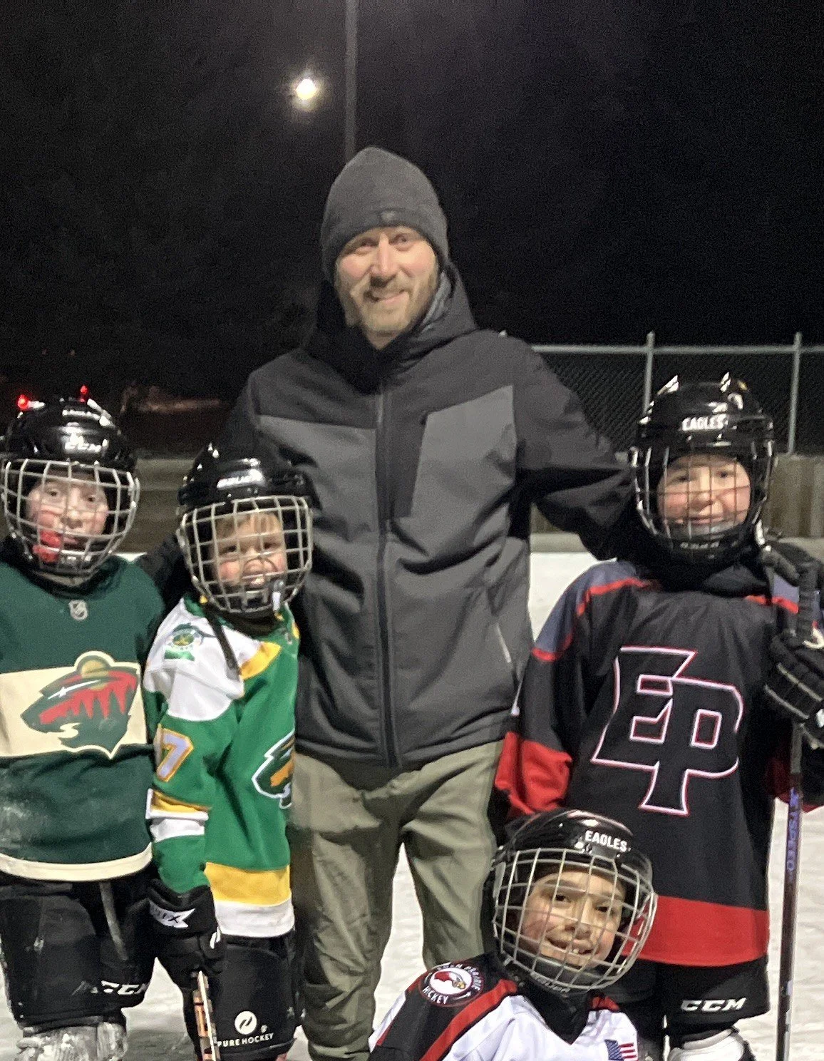 A man in a black jacket and beanie standing with four young children in hockey gear on an ice rink at night, with the moon visible in the sky.