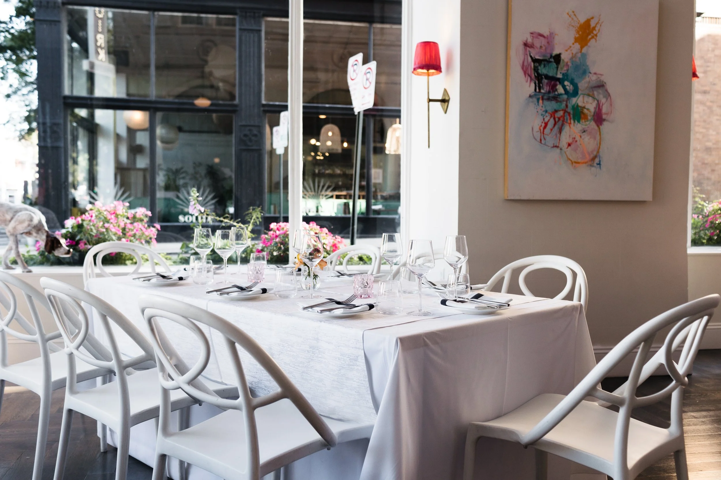 A restaurant dining area with a white tablecloth-covered table set with wine glasses, water glasses, cutlery, and pink glasses. White chairs surround the table. Large windows in the background reveal outdoor planters with pink flowers and a building exterior. The wall features colorful abstract art and a red wall sconce.