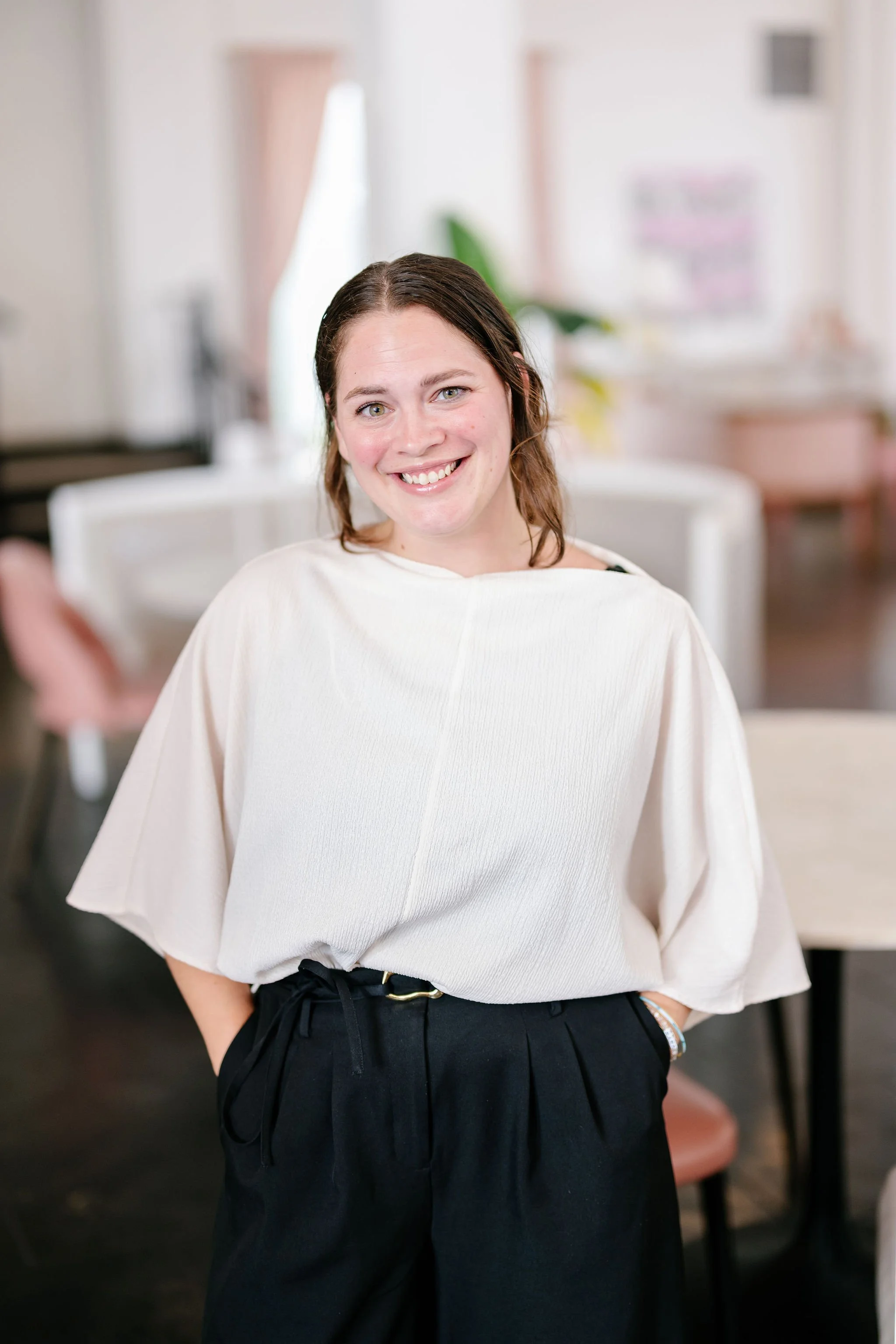 A woman with shoulder-length brown hair smiling, wearing a white top and black pants, standing indoors in a well-lit, cozy room with blurred background furniture and decor.