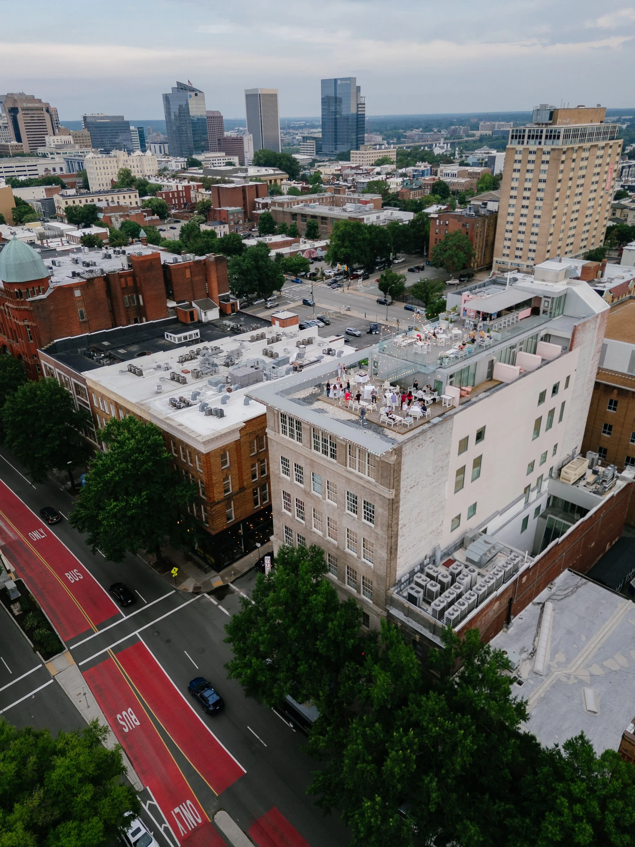 A cityscape view from a high vantage point showing a mix of modern glass skyscrapers and older brick buildings, with a rooftop party area on one building and a red bus lane in the street below.