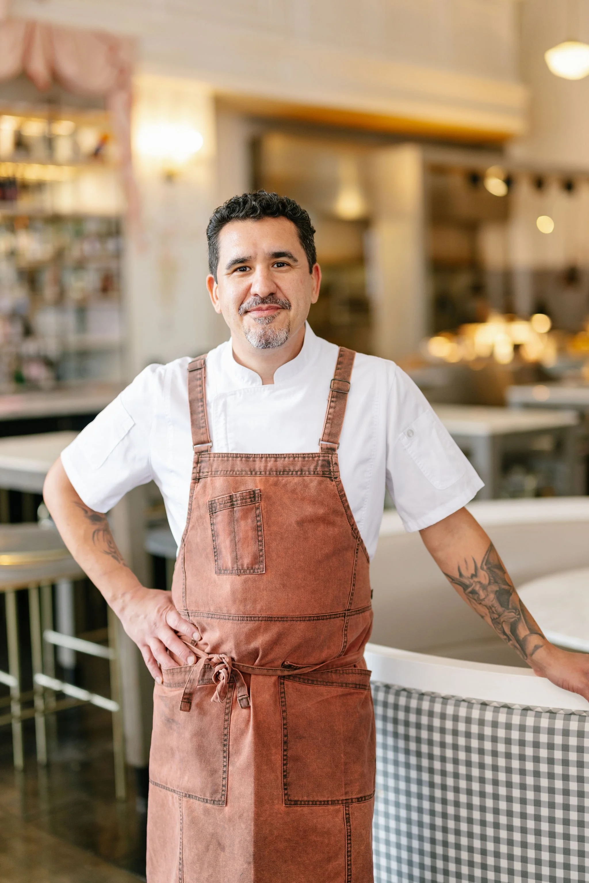 A male chef with black curly hair, a beard, and tattoos on his left arm, standing in a restaurant kitchen, wearing a white chef's coat and a brown apron, smiling.