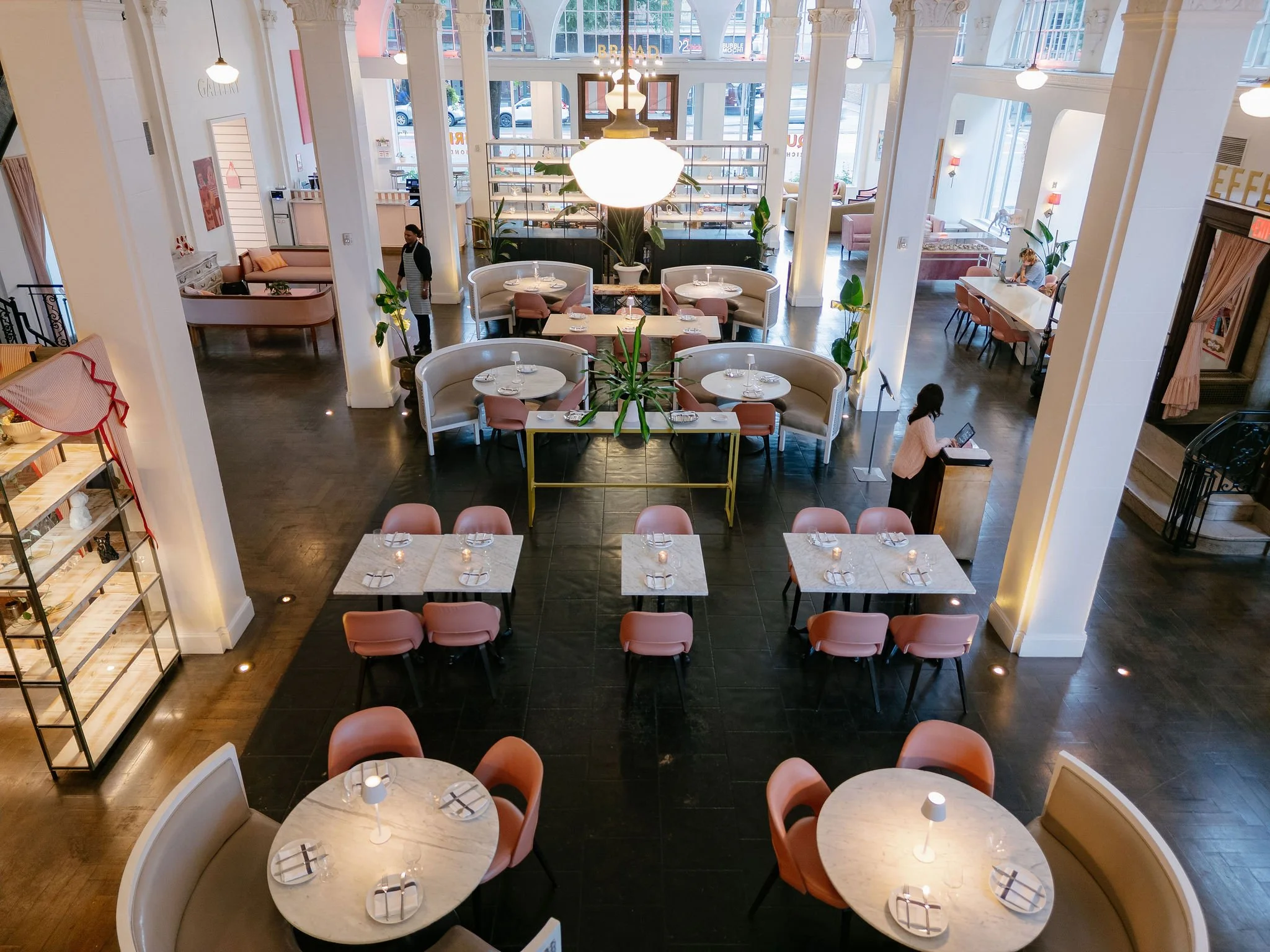 A spacious restaurant with a high ceiling, large windows, and elegant decor. There are round marble tables set with napkins and glasses, and pink upholstered chairs. Large potted plants decorate the area, and a few people are seated or walking around. A prominent chandelier hangs over the center table, and sunlight streams in through the windows.