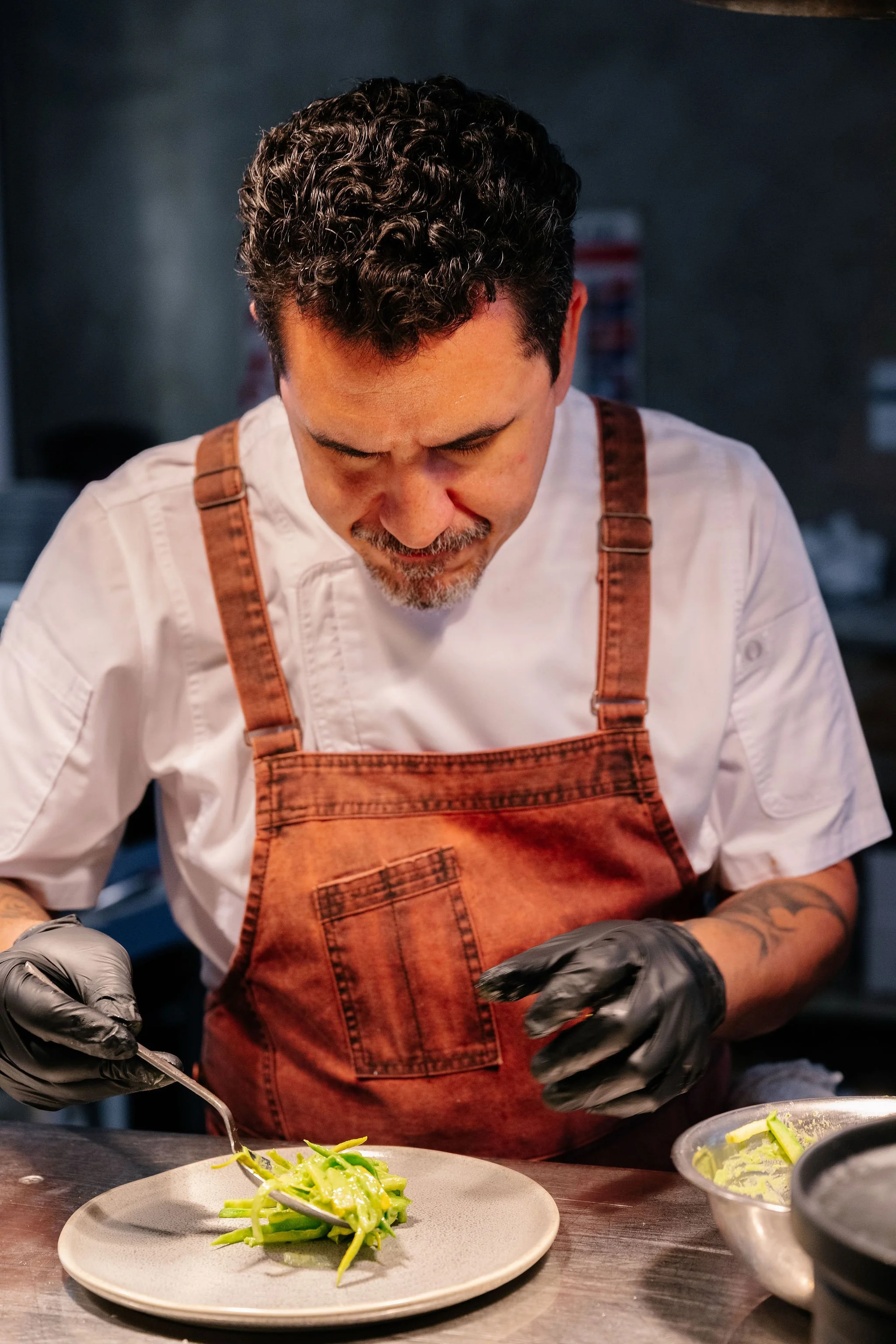 A chef with dark, curly hair, wearing a white shirt and brown apron, is plating or preparing food in a professional kitchen. The image shows a focus on his hands working with food, and a white plate with what appears to be pasta or vegetables in front of him.
